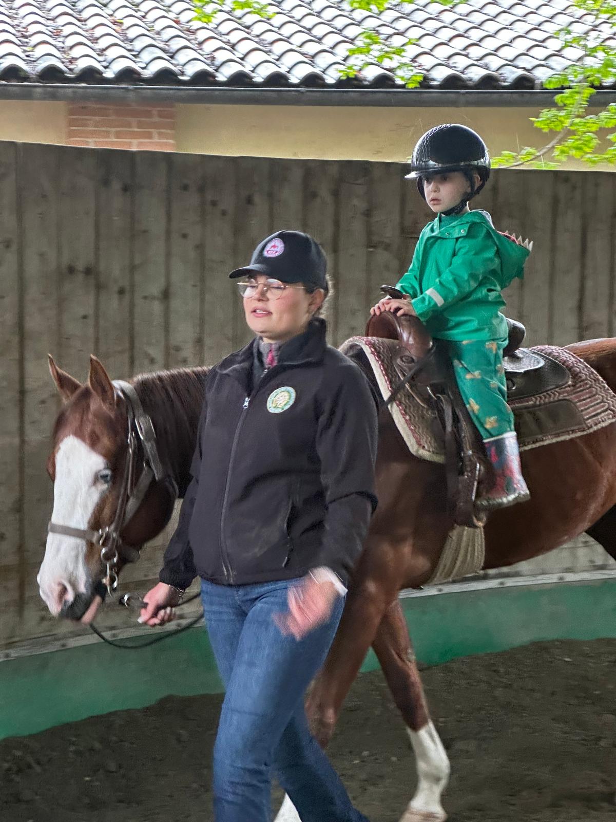 Child riding a horse at the baby farm, guided by an instructor. Safe introduction to farm animals.